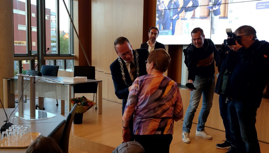 het opspelden van het lintje door burgemeester cees van den bos bij diny jansen. achtergrond de pers met omroep zeeland